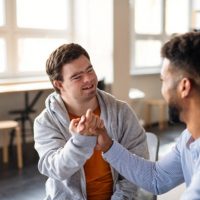 A young happy man with Down syndrome with his mentoring friend celebrating success indoors at school.